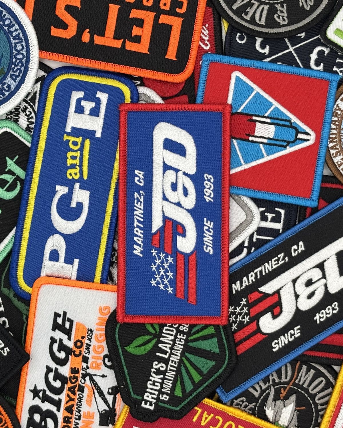 A pile of custom embroidered patches in many shapes and colors from J&D Custom Embroidery, with a J&D 'Since 1993' patch in the center