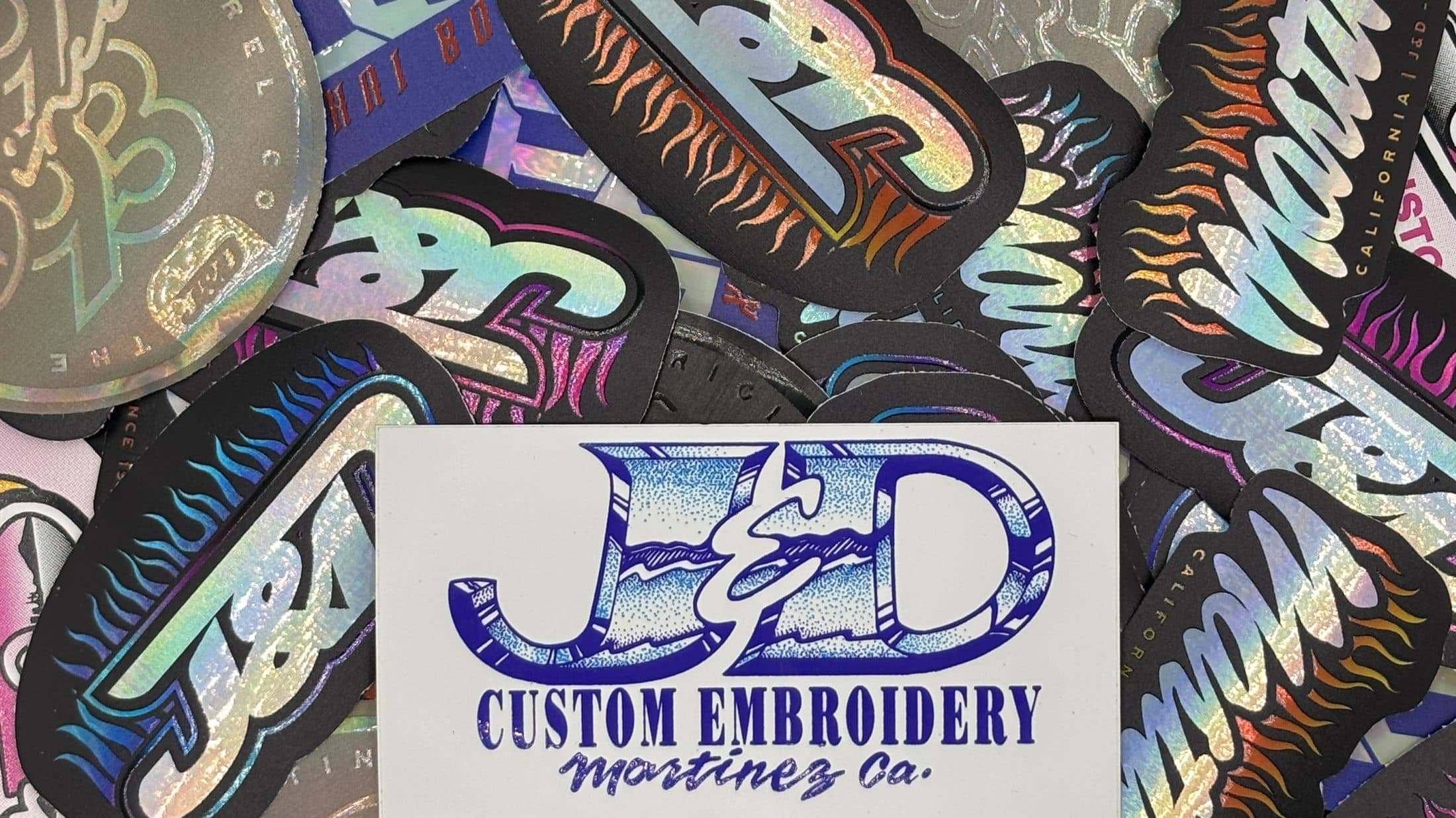 A pile of holographic and matte custom stickers by J&D Custom Embroidery, with a blue J&D Custom Embroidery card in the center